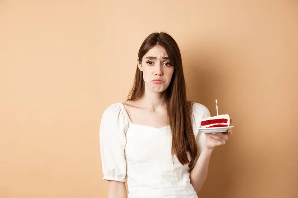 Triste cumpleañera sosteniendo pastel con vela y mirada molesta, sintiéndose sola en su bday, de pie solo sobre fondo beige — Foto de Stock