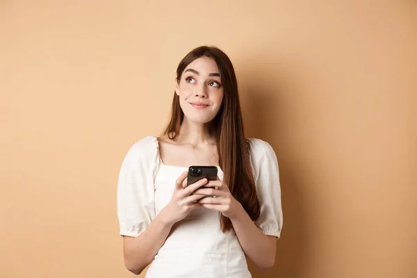 Romantický dívka myšlení s telefonem, dívá stranou a usmívá zasněný, pomocí datování app na smartphone, stojící na béžové pozadí — Stock fotografie