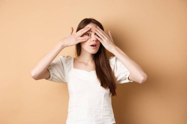 İlginç bir şeye parmak ucuyla bakan meraklı kız şaşkınlığı görmek için sabırsızlanıyor. Kameraya bak, bej arka plan.