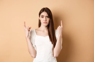 Şüpheci kadın kaşlarını çatıyor ve parmaklarını kaldırıyor, ürünle ilgili şüpheleri var, bej arka planda duruyor.