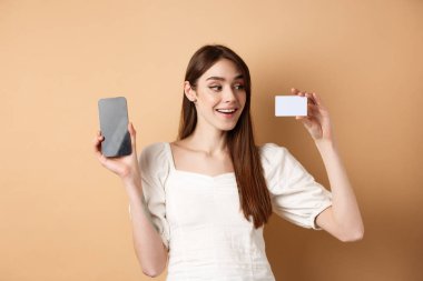 Beyaz bir kadın, mutlu bir gülümsemeyle plastik kredi kartı gösteriyor. Bej arka planda duran boş bir cep telefonu ekranı gösteriyor.