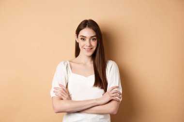 Göğsünde çapraz kollar olan, gülümseyen samimi kız, profesyonel gibi görünüyor, bej arka planda kendine güveniyor.