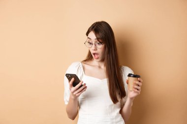 Şaşırmış kız cep telefonundan heyecan verici haberler okuyor, ağzının suyu akıyor, bej arka planda kahve fincanıyla ayakta duruyor.