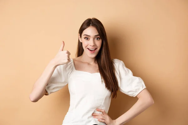 Incrível jovem mulher mostrando polegar para cima, dizer sim e elogiar grande produto, aprovar a escolha, de pé satisfeito em fundo bege — Fotografia de Stock