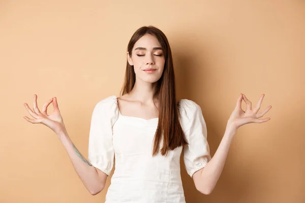 Avslappnad ung kvinna meditera, hålla händerna i yoga zen pose och blunda, känna frid och leende, står på beige bakgrund — Stockfoto