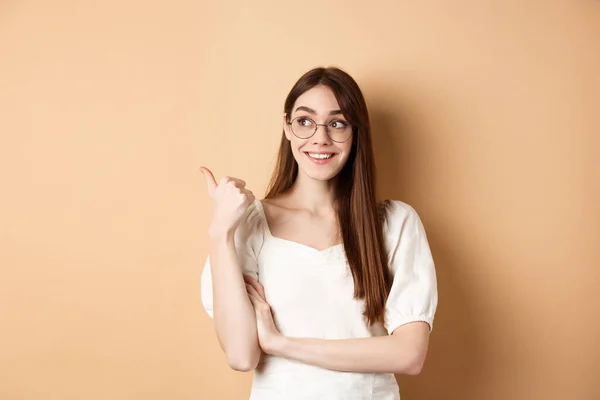 Mujer feliz en gafas sonriendo y apuntando a la izquierda, mostrando el logo en el espacio vacío, de pie sobre fondo beige — Foto de Stock