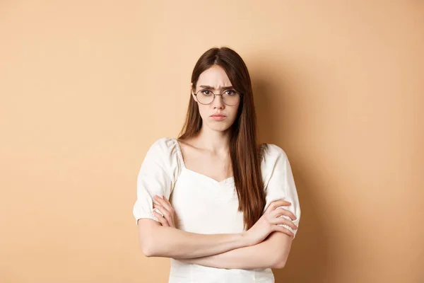 Fille en colère et offensée en lunettes fronçant les sourcils, bras croisés sur la poitrine et boudant, debout défensive sur fond beige — Photo