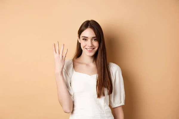 Attraente giovane donna mostra le dita numero quattro, sorridente e guardando fiducioso, in piedi su sfondo beige — Foto Stock