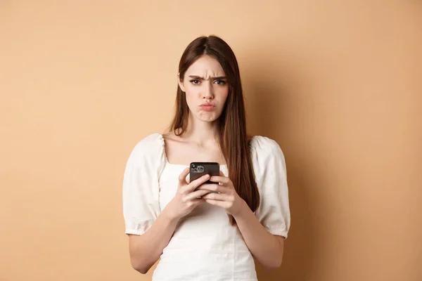 Разочарованная девушка с хмурым смартфоном, грустные губы расстроены, читает плохие новости по телефону, стоит на бежевом фоне — стоковое фото
