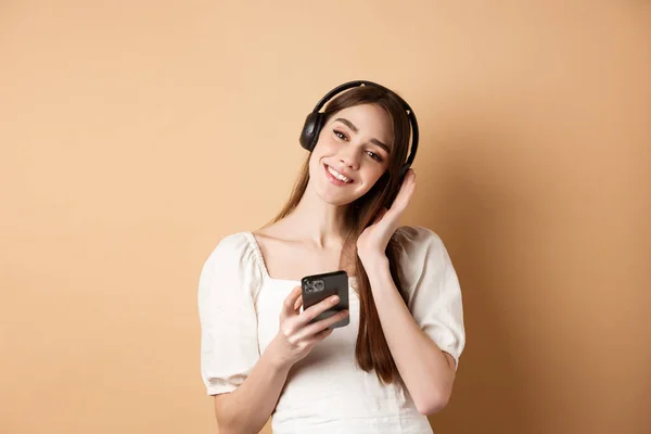 Linda chica sonriente escuchando música en auriculares inalámbricos, usando teléfono inteligente, de pie sobre fondo beige —  Fotos de Stock