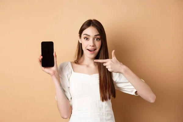 Fröhliche Frau zeigt mit dem Finger auf leeren Telefonbildschirm, sieht aufgeregt aus, steht auf beigem Hintergrund — Stockfoto