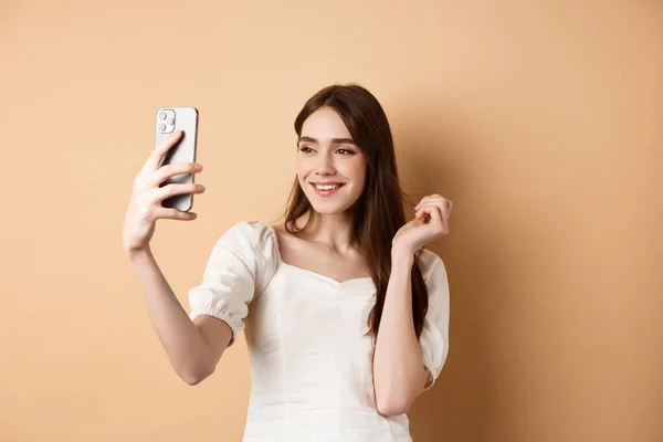 Beautiful woman taking selfie on smartphone, posing for phone photo, standing on beige background — Stock Photo, Image