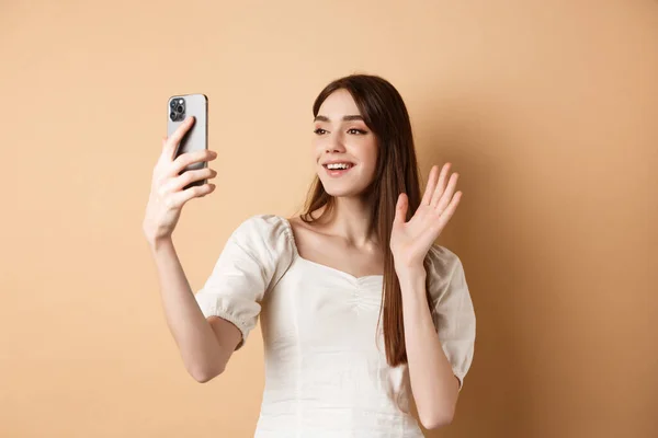 Девушка видео чат по телефону, отказываясь руку на мобильную камеру и говоря привет, разговаривая с другом, улыбаясь и стоя на бежевом фоне — стоковое фото