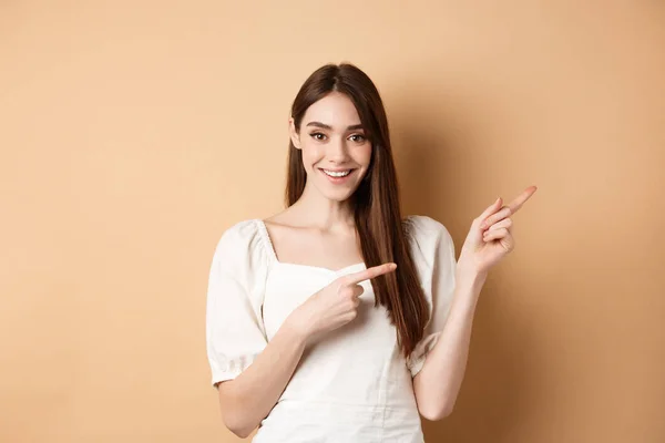 Sorrindo jovem mulher em vestido apontando para o lado e olhando satisfeito, mostrando bom negócio promo, de pé sobre fundo bege — Fotografia de Stock