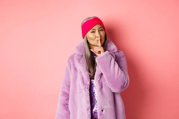 Fashion and shopping concept. Modern chinese senior woman shushing, telling a secret and looking cunning at camera, standing in trendy fake fur coat and headband on pink background — Stock Photo, Image