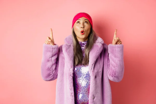 穿着时髦的假毛皮外套、衣着时髦的亚裔老妇人，站在粉红的背景上，凝视着宣传品，抬起头，指指点点 — 图库照片
