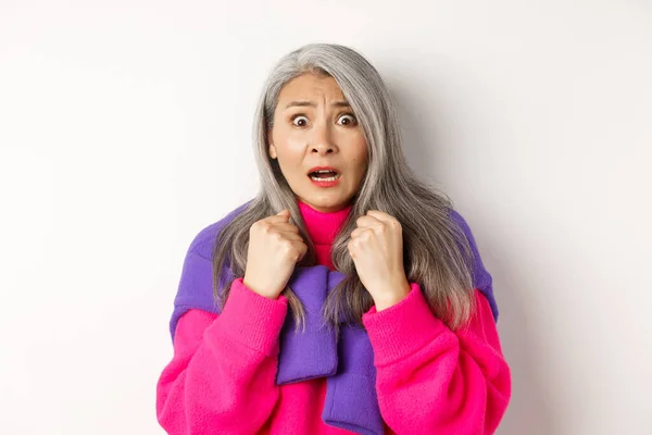 Close up of scared asian senior woman gasping, looking frightened and startled at camera, standing over white background — Stock Photo, Image