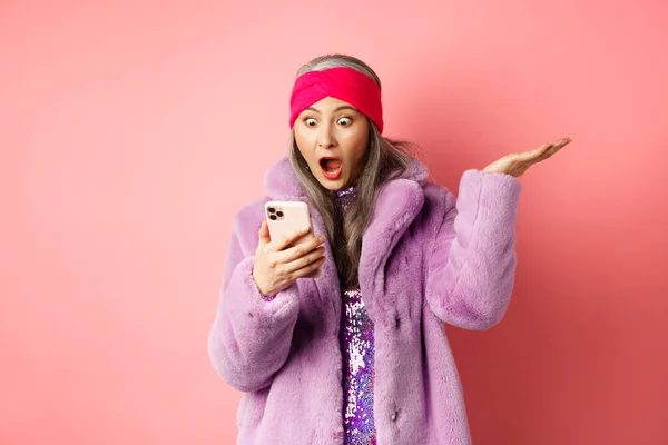 Compras en línea y concepto de moda. Sorprendida y sorprendida asiática mujer mayor mirando la pantalla del teléfono inteligente, leyendo en el teléfono móvil y jadeando, fondo rosa —  Fotos de Stock