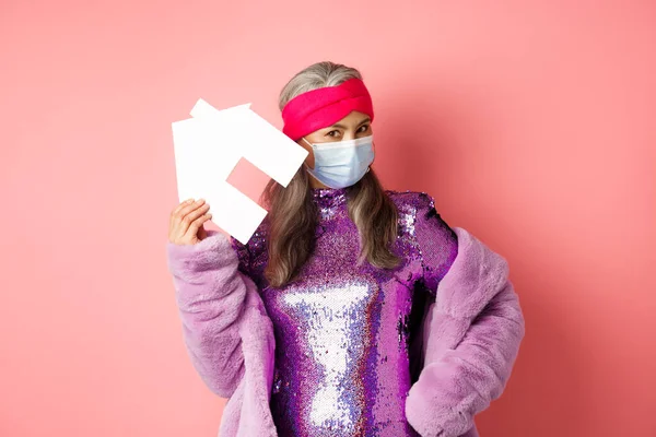 Covid-19 και έννοια ακινήτων. Δροσερό και κομψό ασιατική ηλικιωμένη γυναίκα σε ντίσκο φόρεμα και ιατρική μάσκα που δείχνει χάρτινο σπίτι, στέκεται πάνω από ροζ φόντο — Φωτογραφία Αρχείου