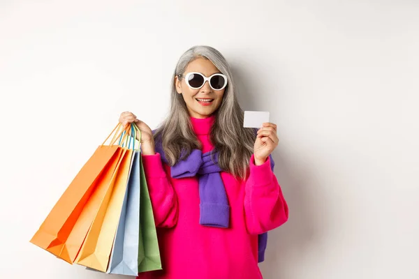 Stilvolle ältere asiatische Frau mit Sonnenbrille, die im Urlaub einkaufen geht, mit Papiertüten und Plastikkreditkarte vor weißem Hintergrund — Stockfoto