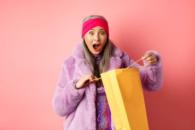 Alışveriş ve moda konsepti. Sürpriz Asyalı büyükanne dükkan çantasını açtı ve kameraya hayret içinde baktı, Anneler Günü hediyesi aldı, pembe arka plan