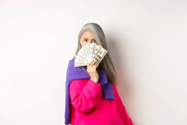Σχέδιο αγορών. Πλούσια και μοντέρνα Κορεάτισσα ηλικιωμένη γυναίκα κρύβεται πίσω από τα χρήματα, κοιτάζοντας την κάμερα, στέκεται πάνω από το λευκό φόντο — Φωτογραφία Αρχείου