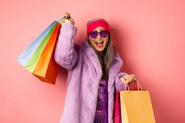 Stilvolle asiatische Seniorin geht einkaufen, trägt trendige Kleidung und Sonnenbrille, hält Einkaufstaschen in der Hand und tanzt fröhlich, rosa Hintergrund — Stockfoto