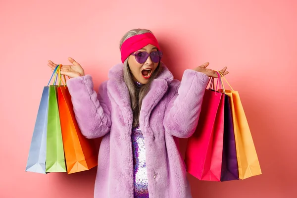 Stilvolle asiatische Seniorin, die einkaufen geht, trendige Kleidung und Sonnenbrille trägt, Einkaufstaschen mit Geschenken in der Hand hält, sich selbst gönnt, rosa Hintergrund — Stockfoto