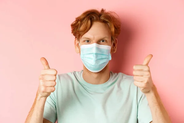 Covid-19 e conceito de pandemia. Bonito cara com cabelo de gengibre bagunçado, usando máscara médica no rosto e mostrando os polegares para cima, de pé sobre fundo rosa — Fotografia de Stock