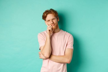 Elinde kızıl saçlar, kameraya yorgun ve keyifsiz bakan, turkuaz arka planda tişörtle duran sıkkın yakışıklı adam.