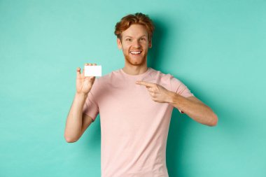 Sakallı ve kızıl saçlı çekici yetişkin adam plastik kredi kartını işaret ediyor, kameraya gülümsüyor, turkuaz arka planda duruyor.