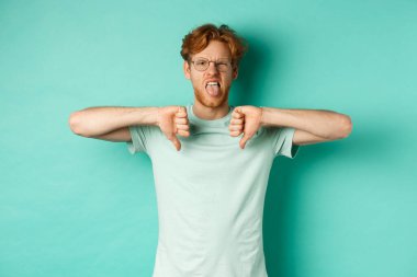 Tişört ve gözlüklü yargılayıcı kızıl saçlı erkek model, dil gösteriyor ve hoşnutsuz, hayal kırıklığını ve hoşnutsuzluğu ifade etmek için başparmaklarını öne eğiyor, turkuaz arka plan