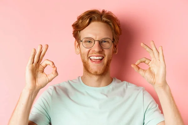 Retrato headshot de jovem atraente com cabelo vermelho confuso e barba, vestindo óculos, sorrindo com dentes brancos e mostrando Ok sinais em aprovação, louvar algo bom — Fotografia de Stock