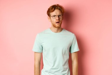 Gözlüklü, kafası karışık, ağzı açık, kameraya bakan, hiçbir şey anlamayan, pembe arka planda duran yakışıklı kızıl adamın portresi.
