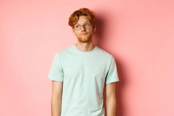 Imagen del joven pelirrojo con barba, vistiendo camiseta y gafas, mirando a la izquierda con la cara aburrida y tranquila, de pie sobre fondo rosa —  Fotos de Stock