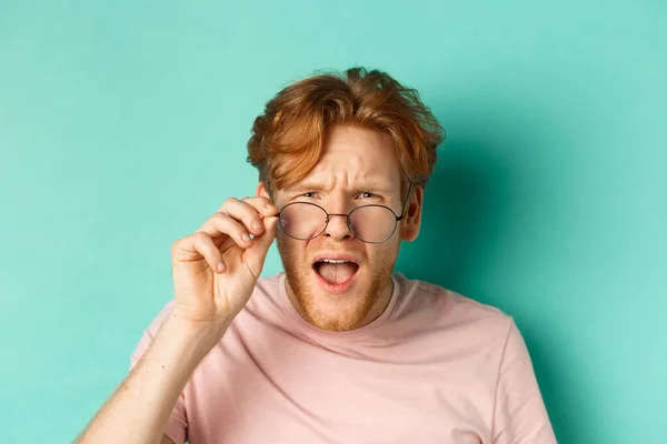 Close up portrait of redhead guy take-off glasses and looking confused at something strange, standing shocked over turquoise background — Stock Photo, Image
