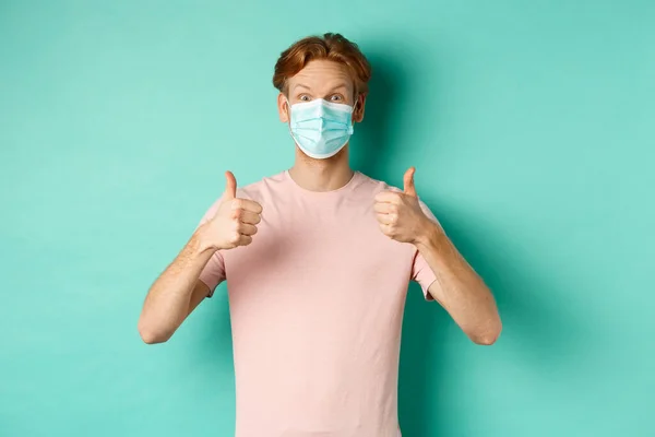 Covid-19, pandemic and lifestyle concept. Cheerful redhead guy in medical mask showing thumbs up in approval, like and praise product, standing over turquoise background — Stock Photo, Image
