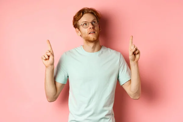Dromerige jongeman met rood haar en baard wijzend vingers omhoog, op zoek pensive aan de bovenkant, staande tegen roze achtergrond — Stockfoto