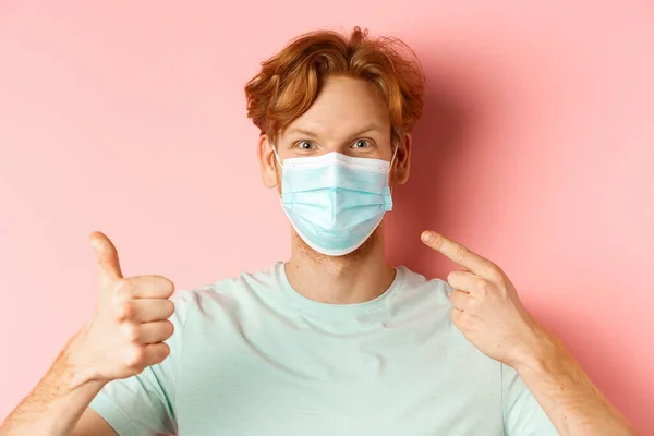 Covid-19 e conceito de pandemia. Bonito ruivo cara apontando dedo na máscara facial e mostrando polegares para cima, usando medidas de coronavírus, de pé sobre fundo rosa — Fotografia de Stock