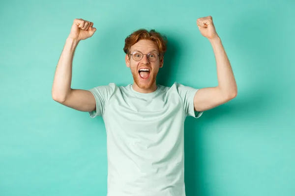 Joven pelirrojo sintiéndose como campeón, levantando las manos en el gesto de la bomba de puño y gritando sí con alegría, ganando el premio, triunfando del éxito, de pie sobre fondo de menta — Foto de Stock