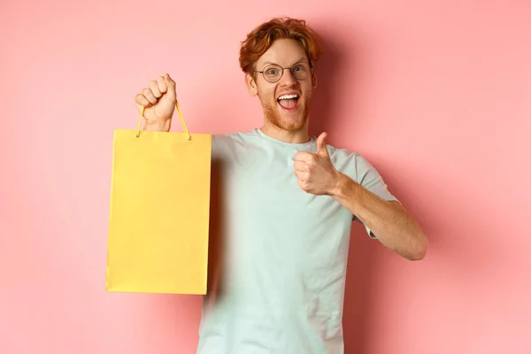 Hombre pelirrojo alegre en camiseta y gafas señalando con el dedo a la bolsa de la compra, mostrando tienda con descuentos, de pie sobre fondo rosa —  Fotos de Stock
