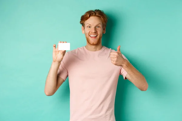 Wesoły klient banku mężczyzna w t-shirt pokazujący kciuk w górę i plastikową kartę kredytową, uśmiechnięty zadowolony przed kamerą, stojący nad turkusowym tle — Zdjęcie stockowe