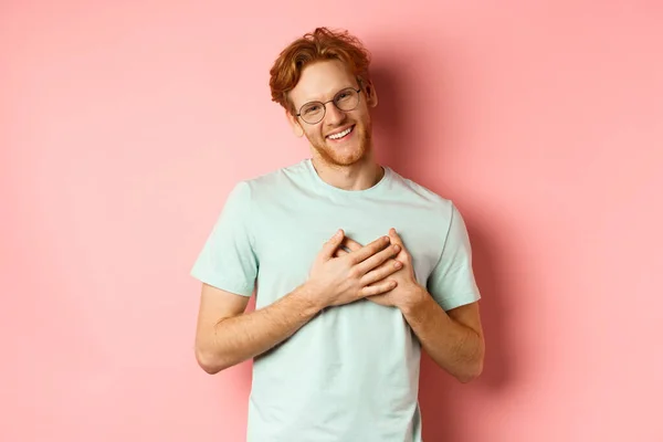 Attractive young man with ginger hair, holding hands on heart and smiling grateful, saying thank you, express gratitude, standing over pink background