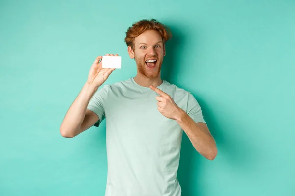 Koncepcja zakupów. Przystojny rudzielec w t-shircie z plastikową kartą kredytową i uśmiechnięty, stojący na turkusowym tle — Zdjęcie stockowe