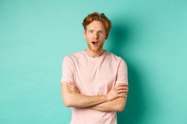 Dedikoduları dinleyen, kameraya hayran ve ilgili görünen, turkuaz arka planda günlük tişörtle dikilen ilginç kızıl saçlı adam.