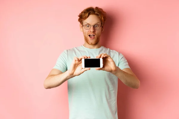 Hombre pelirrojo asombrado jadeando y mirando con asombro el teléfono, mostrando la pantalla del teléfono inteligente en blanco horizontalmente, de pie sobre el fondo rosa — Foto de Stock