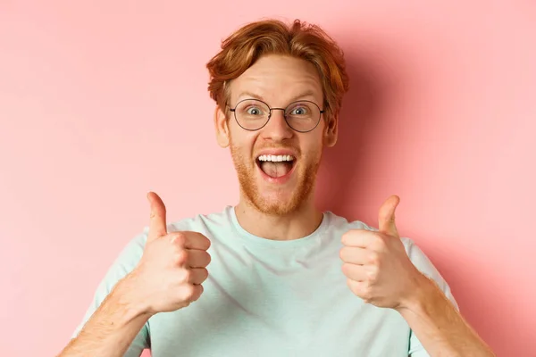 Gesicht eines glücklichen rothaarigen Mannes in Brille und T-Shirt, Daumen nach oben zeigend und aufgeregt guckend, billigend und lobend coole Promotion, vor rosa Hintergrund stehend — Stockfoto