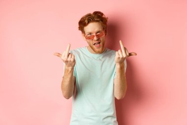 Arrogant and careless redhead man in sunglasses dont give a fuck, showing middle fingers at camera and frowning, standing over pink background
