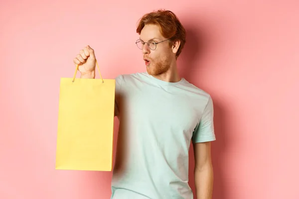 Handsome young man buying presents, holding shopping bag and looking amused, standing over pink background