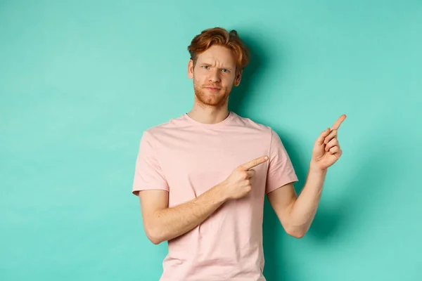 Sceptische en twijfelachtige roodharige man in t-shirt wijzende vingers op de rechter bovenhoek, tonen promo aanbod met ontevreden gezicht, staande over turquoise achtergrond — Stockfoto